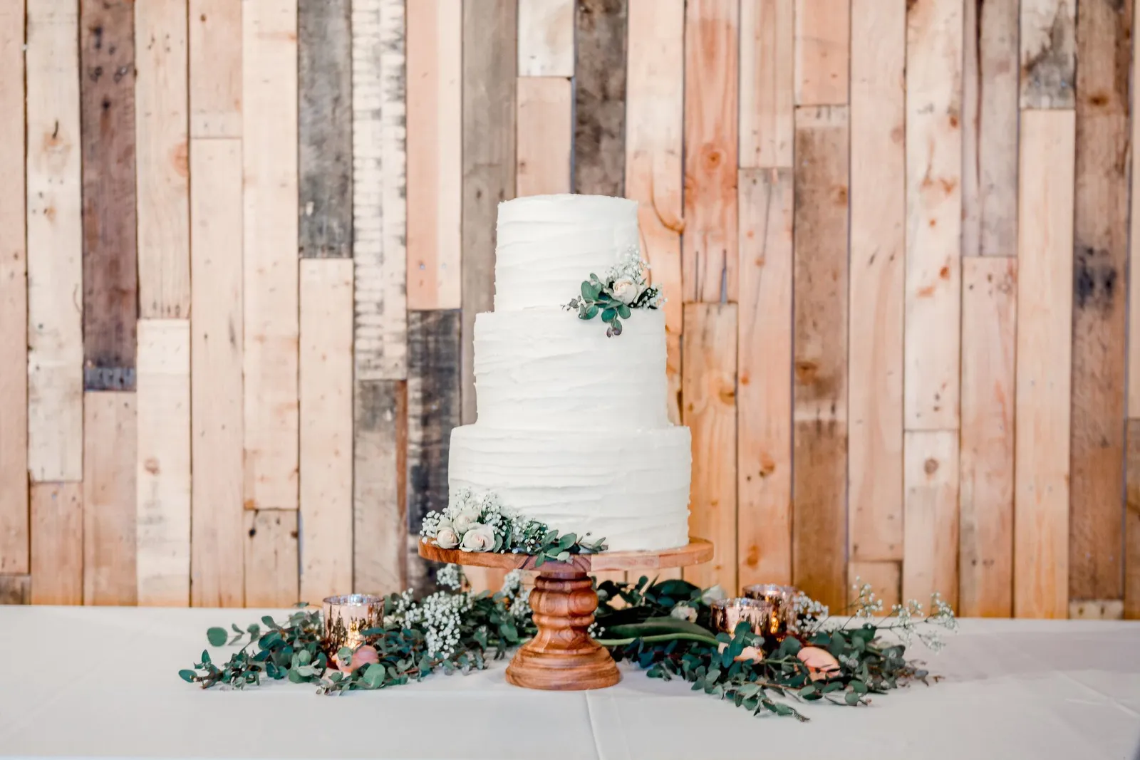 Three-tier white wedding cake with greenery and floral accents on a wooden stand